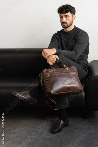 A professional male model in charcoal knitwear and tailored trousers sits on a black leather sofa holding a premium brown leather briefcase.