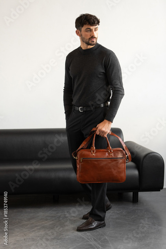 A professional man in a charcoal crewneck sweater and tailored trousers holds a light brown briefcase standing in a minimalist studio.