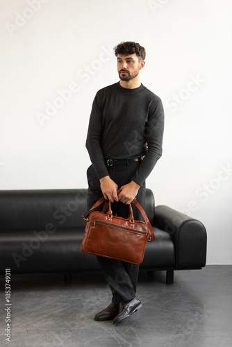 A professional man in a charcoal crewneck sweater and tailored trousers holds a light brown briefcase standing in a minimalist studio.