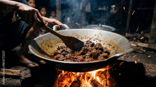 Person stirring meat in a large wok over an open wood fire, creating sparks and smoke at night
