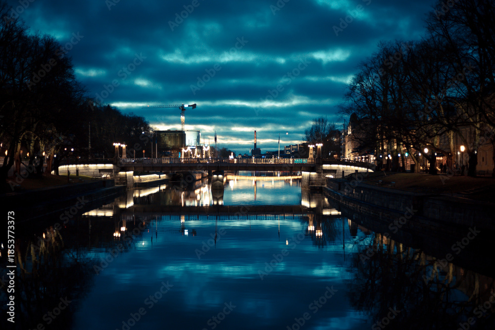 Obraz premium Photo of a night city located along a river, with bright city lights reflecting on the water surface. The image captures an atmospheric urban night scene, ideal for travel