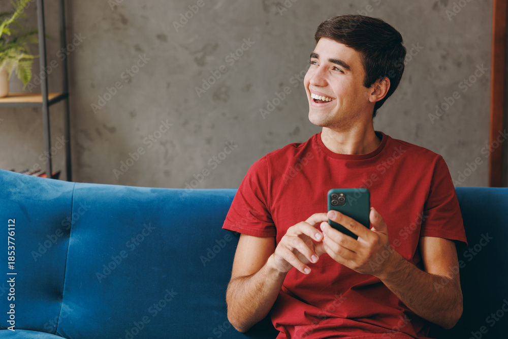 Fototapeta premium Young cheery man wear red t-shirt look aside hold use mobile cell phone sit on blue sofa stay at home hotel flat rest relax spend free spare time in living room indoor grey wall People lounge concept