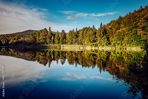 View of the serene lake mirroring the vivid autumn foliage and cerulean sky above, framed by distant hills, Plitvicka Jezera, Lika-Senj County, Croatia.