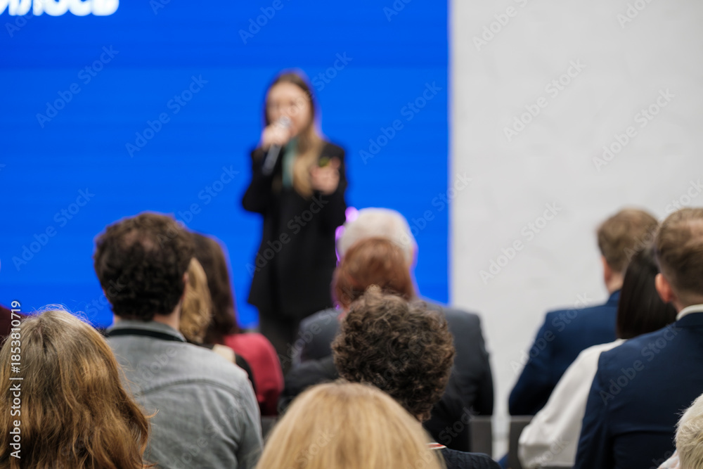 Obraz premium Speaker on stage with microphone addresses attendees. Blue backdrop creates vibrant conference setting. Audience sits attentive.