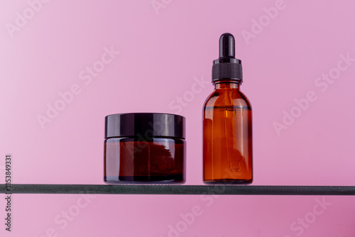 Amber glass skincare bottles and jar with black lids on a shelf against pink background