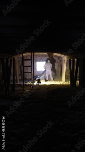 Worker spreads loose fill insulation in loft with hose and rafters. White suit, brick window, ladder, warm work light, low key lighting, vertical frame, documentary mood.