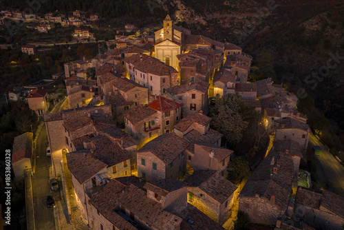 Aerial view of a medieval village glows warmly against the night, the church standing tall amidst the tightly packed buildings, Cottanello, Lazio, Italy.