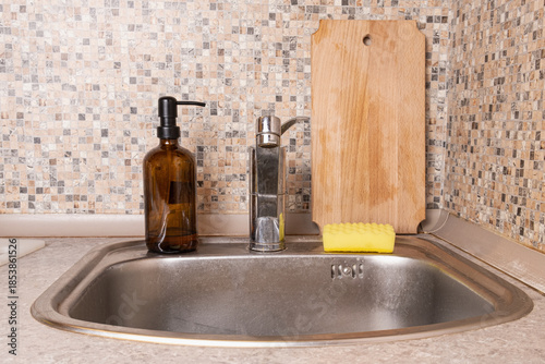 Kitchen sink with refillable soap dispenser, sponge, and wooden cutting board against mosaic backsplash, illustrating everyday dishwashing routine