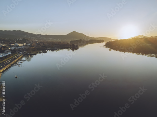 Aerial view of the sun kissing the placid waters near the iconic bridge and marina, casting long shadows over Vila Nova de Cerveira, Viana do Castelo District, Portugal.