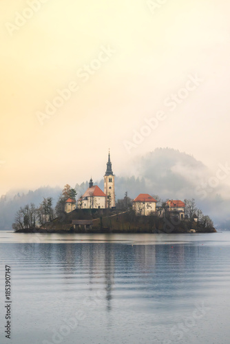 historic church and buildings on misty Bled island surrounded by calm lake water in soft morning light
