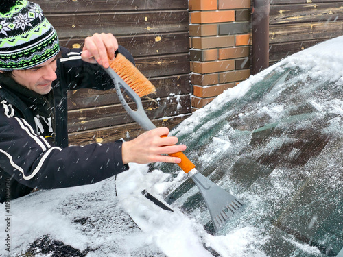 Man scraping snow from car windshield with an ice scraper and brush. Winter concept for cold weather, vehicle maintenance, and daily commute preparation.