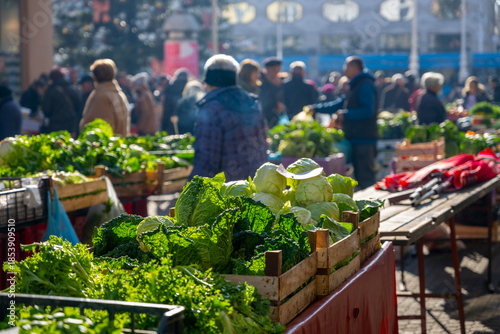 Fresh vegetables displayed at outdoor market with diverse crowd shopping in bright daylight