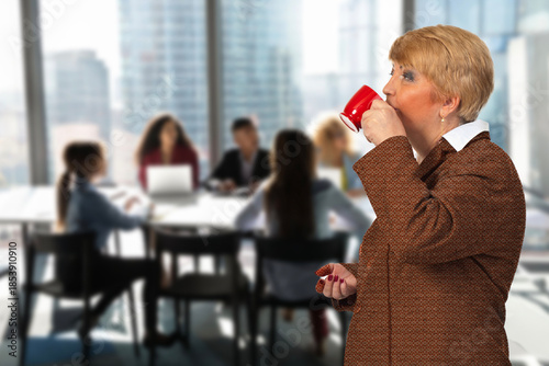 A woman in a red sweater is drinking coffee in front of a group of people. The scene is set in a conference room with a table and chairs. The woman is the only one drinking coffee