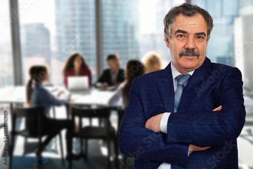 A man in a suit stands in front of a group of people in a conference room. He is wearing a tie and has his arms crossed. The scene suggests a professional setting