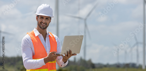 Smiling male engineer in safety vest and helmet using laptop at wind farm, representing renewable energy technology, field operations, sustainable development.