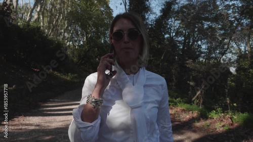 A blonde businesswoman talks on the phone during her lunch break in a park. Modern work-life balance scene combining career, communication, productivity, and outdoor relaxation.