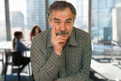 A man with a beard and mustache is sitting at a table with his hands on his face. He looks tired and is staring off into the distance. The scene takes place in a room with a few chairs and a desk