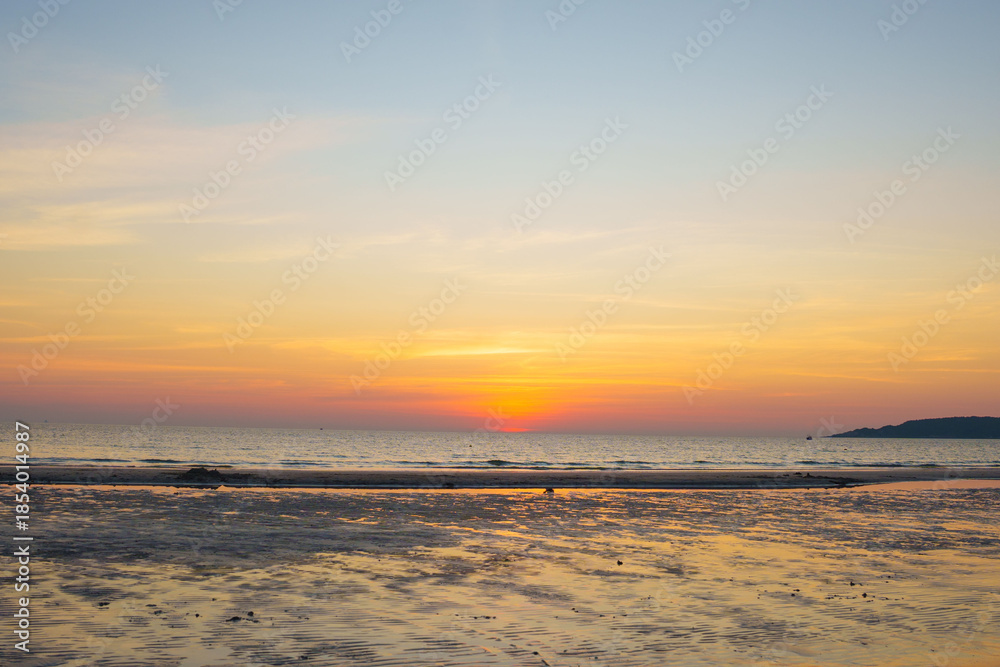 Fototapeta premium Golden sunset over a calm ocean with the sun touching the horizon, warm orange sky reflected on wet sand, creating a peaceful and serene seaside landscape.