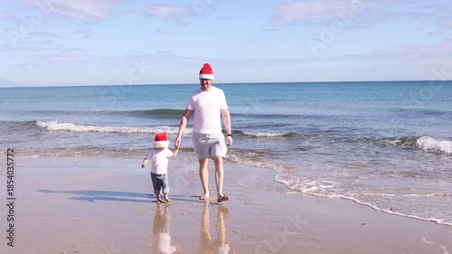 Father and his little son wearing Santa hats walking on a beach near sea. Christmas concept.