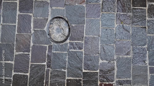 Top-down view of a stone-paved sidewalk in a small town as rain falls steadily, accompanied by distant thunder, capturing the raw atmosphere of a passing storm.