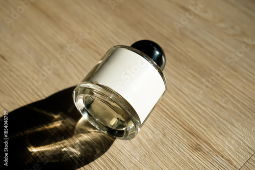 Clear glass perfume bottle with blank label lying on wooden floor, sunlight casting strong shadow