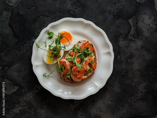 Delicious breakfast, brunch, tapas, snack - boiled egg and avocado, shrimp sandwich on a dark background, top view