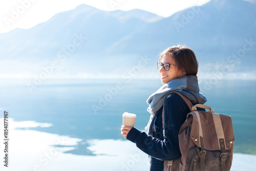 Happy woman traveling by mountain lake, drinking coffee. Traveler enjoying winter sea beach, sabbatical trip for mental health, adventure, relaxing. Girl with backpack on coast. Solo female tourism