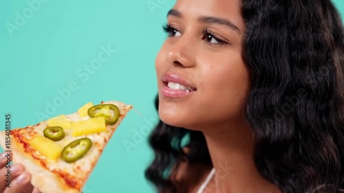 Joyful moment of a woman savoring a pizza slice with pineapple and jalapenos against a turquoise background