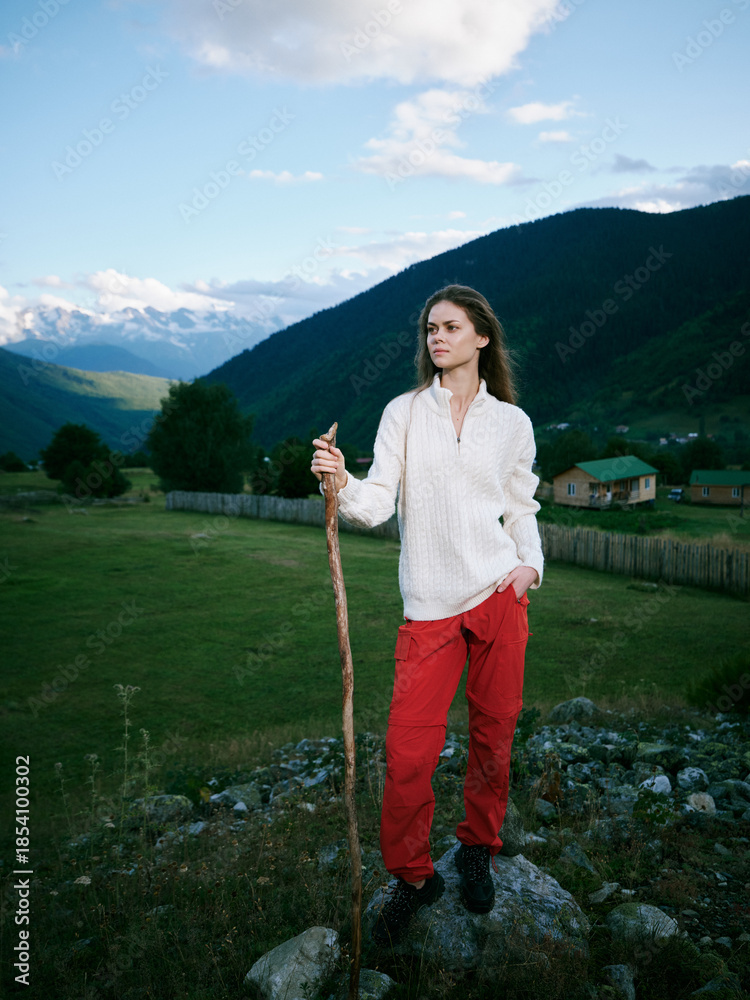 Naklejka premium Woman standing on rocky hill with walking stick in a green valley, mountains in distance, white sweater and red pants, exploring nature and open countryside