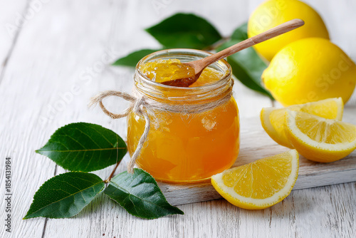 Homemade lemon jam in glass jar with fresh lemons and green leaves on white wooden background, natural citrus preserve for breakfast, desserts, baking, organic food concept, rustic flat lay