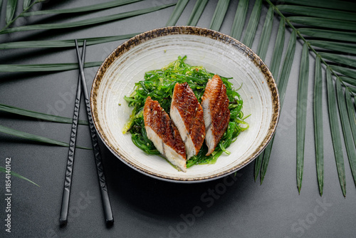 Grilled Eel Slices Over Fresh Green Seaweed Salad. Three pieces of perfectly glazed grilled eel rest atop a bed of bright green Japanese seaweed salad, served in a ceramic bowl.