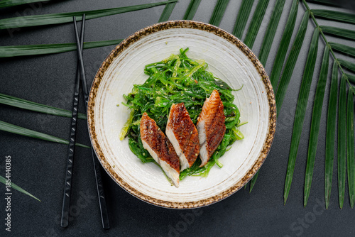 Grilled Eel Slices Over Fresh Green Seaweed Salad. Three pieces of perfectly glazed grilled eel rest atop a bed of bright green Japanese seaweed salad, served in a ceramic bowl.