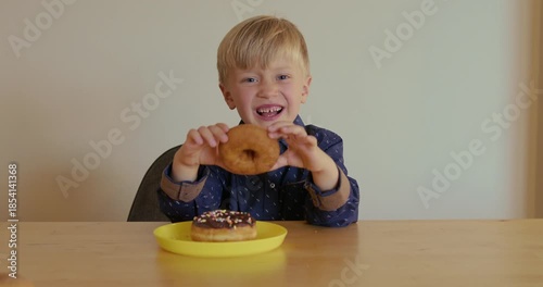 A joyful young boy with two chocolate donuts on a plate lifts one and looks through the hole of the donut. Enjoyment of sweets