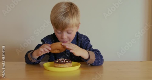 The boy is holding two chocolate donuts with colorful sprinkles and takes a bite of one, then the other.