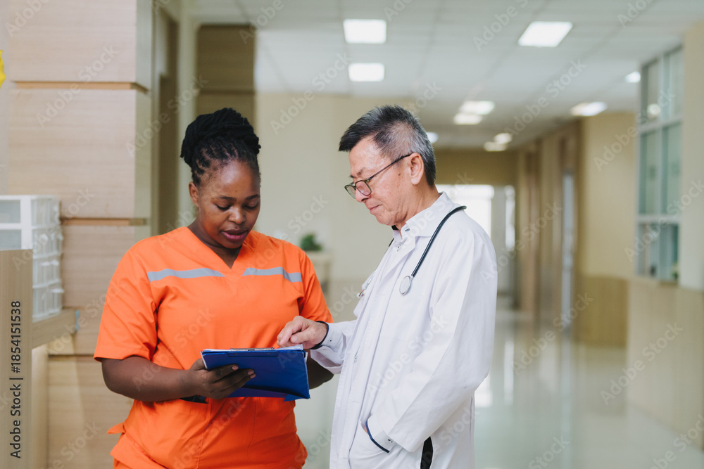 Obraz premium A group of healthcare professionals stands in a hospital corridor, discussing medical documents. Two doctors in white coats and a nurse in bright orange scrubs are engaged in a serious conversation ab