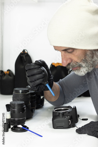A man in a white beanie and black gloves uses a rubber air blower to clean a camera sensor. Multiple lenses and cleaning tools are neatly arranged on the white desk in front of him.