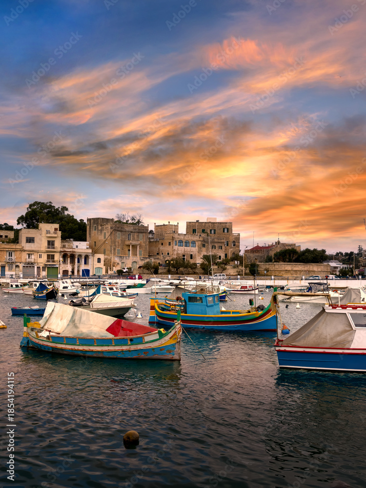 Fototapeta premium View of Birzebbuga's small harbor with numerous moored boats at sunset ; rows of houses appear in the background.