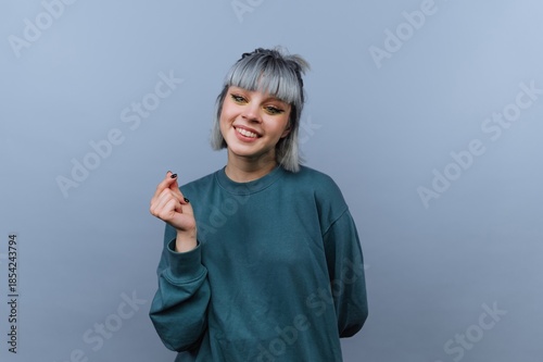 Young woman smiles and poses with a hand gesture in a studio with a plain gray background during bright indoor lighting