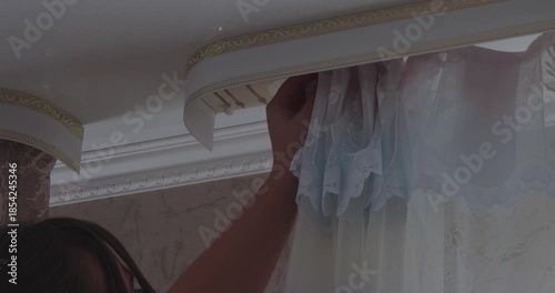 A woman's hand hangs beautiful tulle and lace curtains on a classic curtain rod. The woman is busy with household chores and decorating the room with elegant curtains to create a cozy atmosphere.