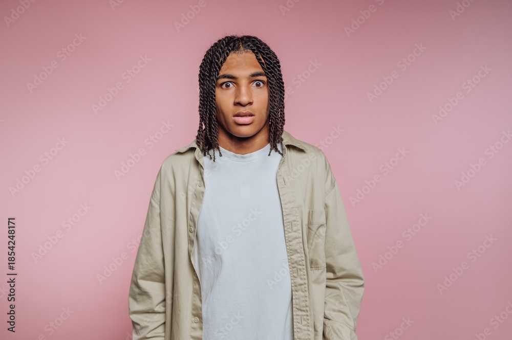 Fototapeta premium Person with braided hair stands against a pink background looking surprised or confused while wearing a casual outfit