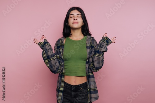 Young woman poses with closed eyes and relaxed posture against a plain pink background, wearing a green top and a plaid shirt