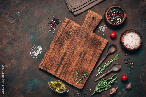 An empty wooden cutting board with various spices, herbs, and seasonings on a dark rusty grunge background. A culinary backdrop with cooking ingredients. Top view, flat lay, copy space.