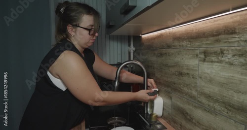 Focused woman wearing glasses doing daily household chores, cleaning dishes with a sponge and soap under running tap water in a modern kitchen after dinner in the evening.