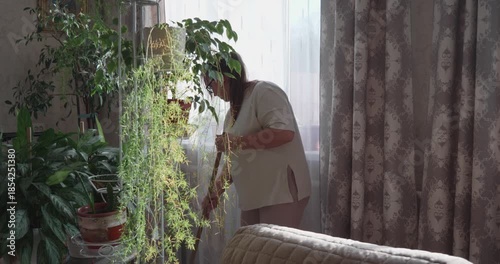 A diligent woman in glasses, busy with her daily chores, sweeps the floor with a broom in a cozy room filled with houseplants. The woman maintains cleanliness and order in her comfortable home.