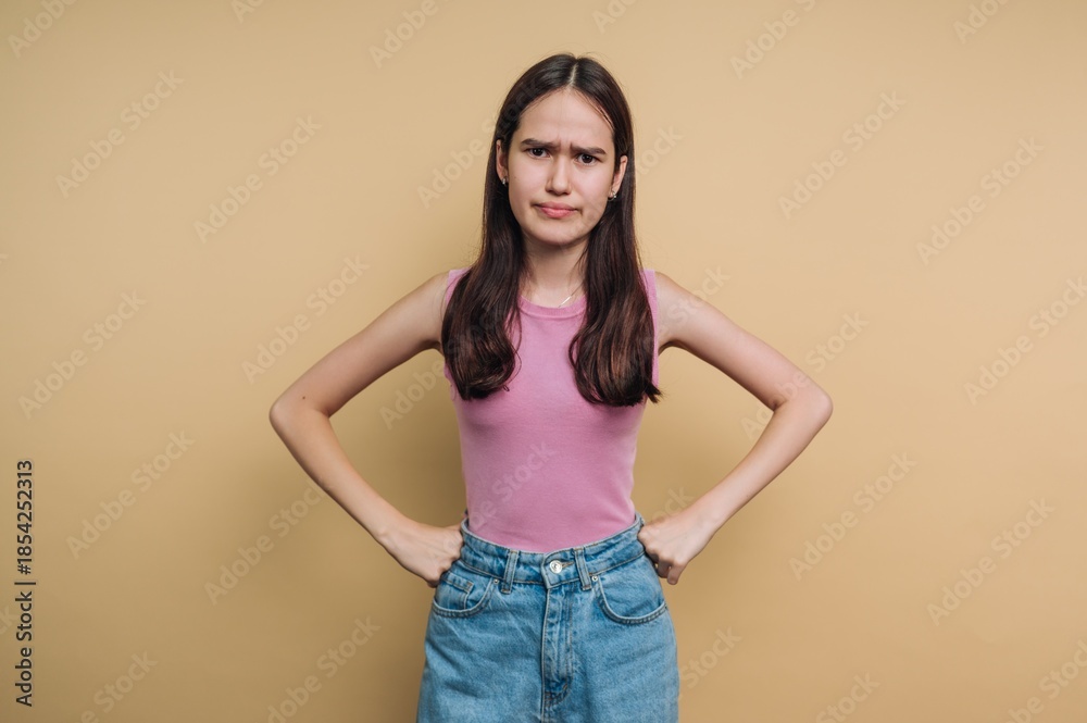 Obraz premium Expression of concern from a young girl with hands on hips in front of a plain background during daytime