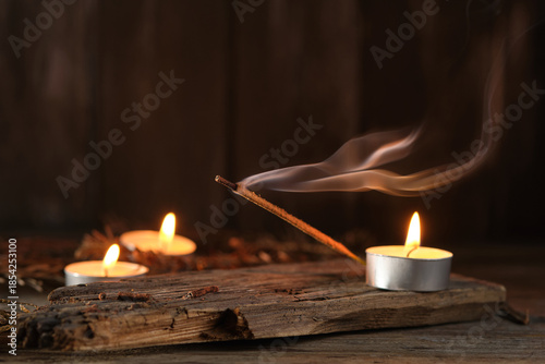 Incense Stick Releases Smoke Next to Candles on Wooden Surface at Night