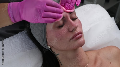 A woman receives a facial cleansing treatment with a white mask applied and removed by an esthetician wearing pink gloves.