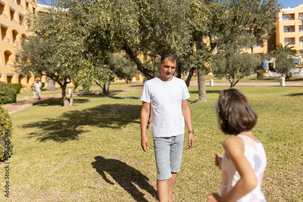 Obraz premium Father watches his daughter run in blur on lawn next to hotel among olive trees.