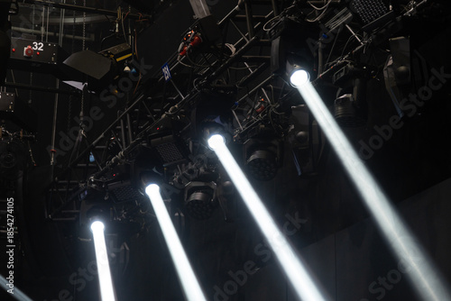 Stage lighting equipment illuminates a dark venue, with multiple moving head lights emitting strong white beams, all mounted on a robust metal truss structure