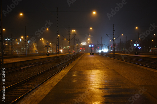 an empty railway station platform, with wet pavement reflecting warm streetlights and fog softening the distant city lights
